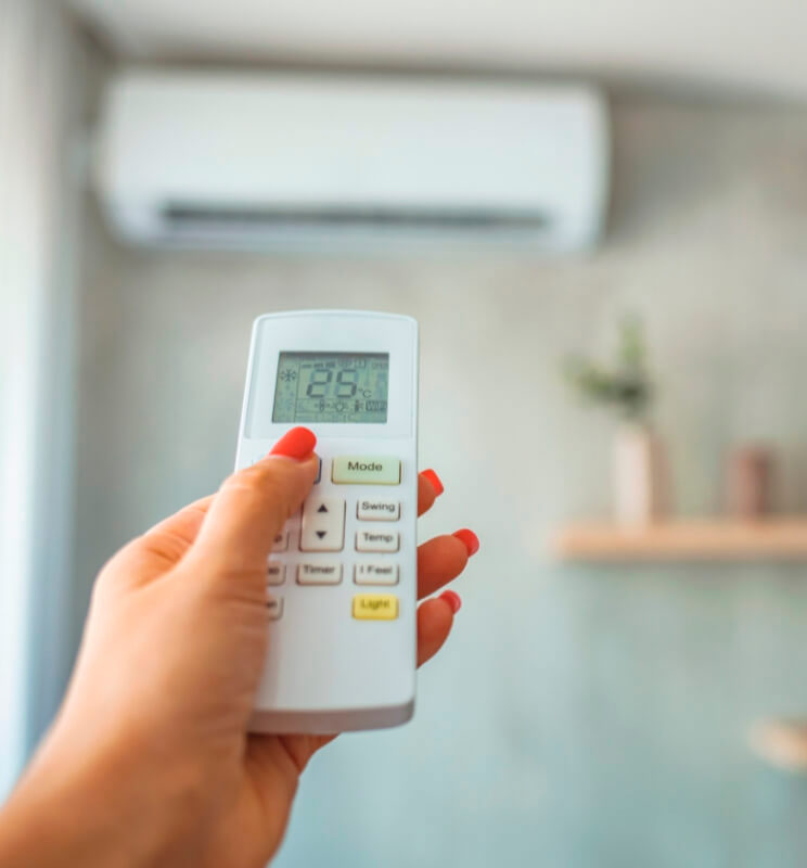 A hand holding an air conditioner remote, displaying a temperature of 26°C, with a unit visible on the wall in a modern room.