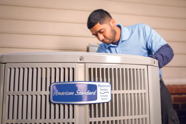 An HVAC technician performs service on an outdoor American Standard Platinum Series variable-speed air conditioning unit.