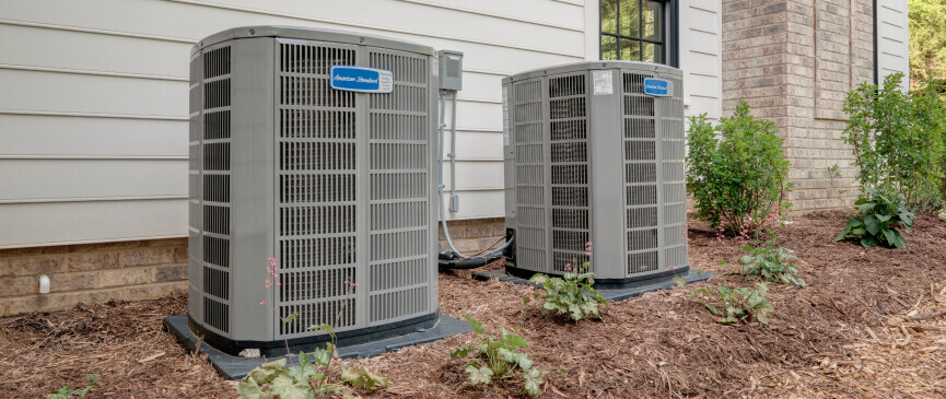 Two American Standard outdoor air conditioning units are neatly installed within a landscaped mulch bed next to a residential home.