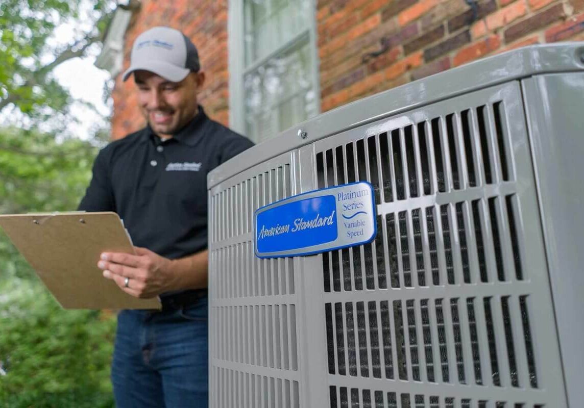 A professional HVAC technician smiles while reviewing paperwork next to a new American Standard Platinum Series variable-speed air conditioner.